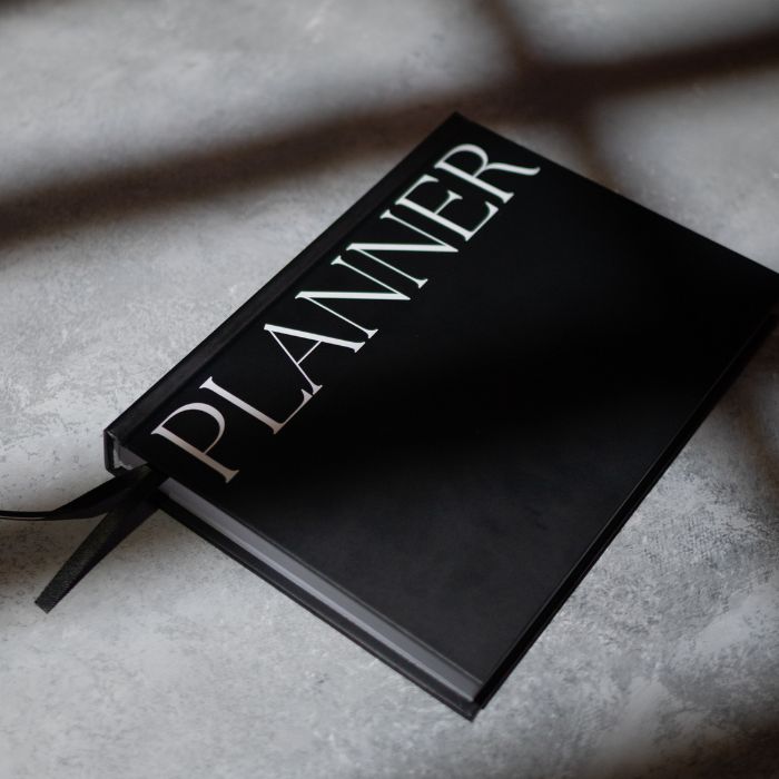 A black hardcover planner with the word 'PLANNER' in large white serif letters on the cover, resting on a textured light gray surface with dramatic shadows.