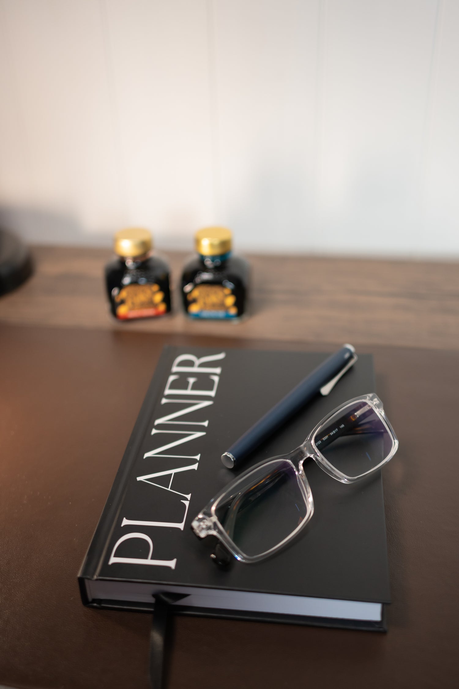 A black hardcover planner with large white 'PLANNER' text, placed on a wooden desk with a dark pen and clear-framed eyeglasses resting on top. Two small bottles are blurred in the background against a light wall.