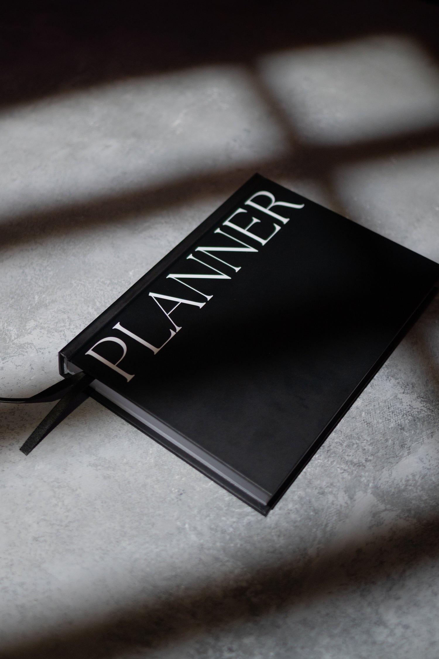 A black hardcover planner with the word 'PLANNER' in large white serif letters, photographed on a grey textured surface with dramatic window-box shadows.