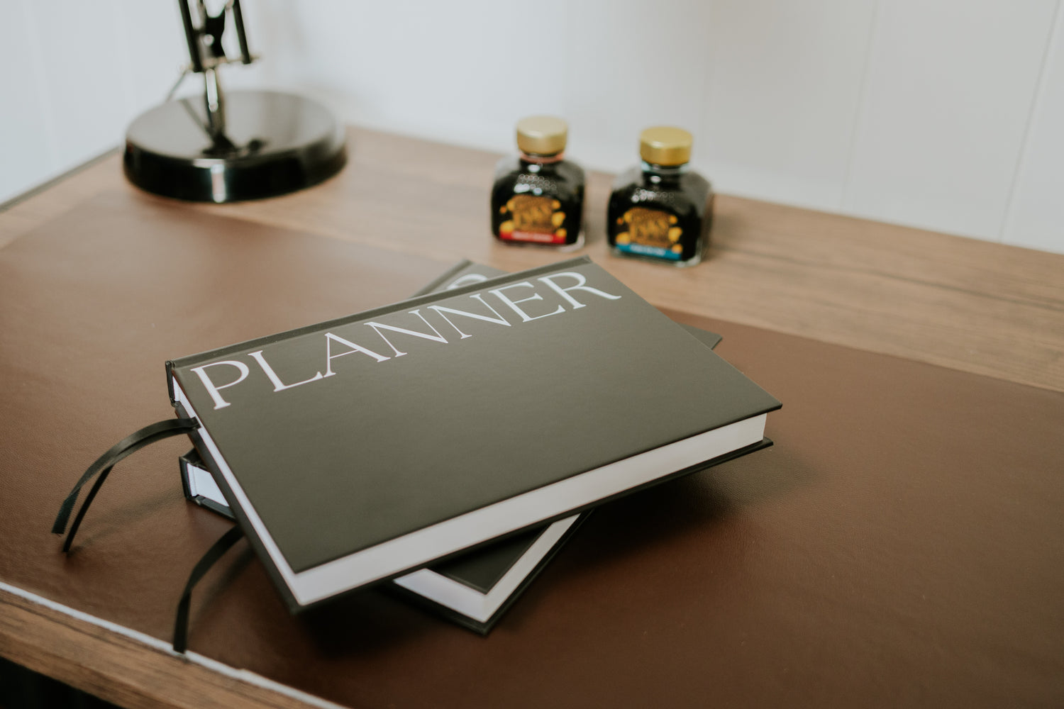 A hardcover planner with the word 'PLANNER' in large white letters on a dark cover, resting on a wooden desk. Two small decorative bottles are blurred in the background near a lamp.