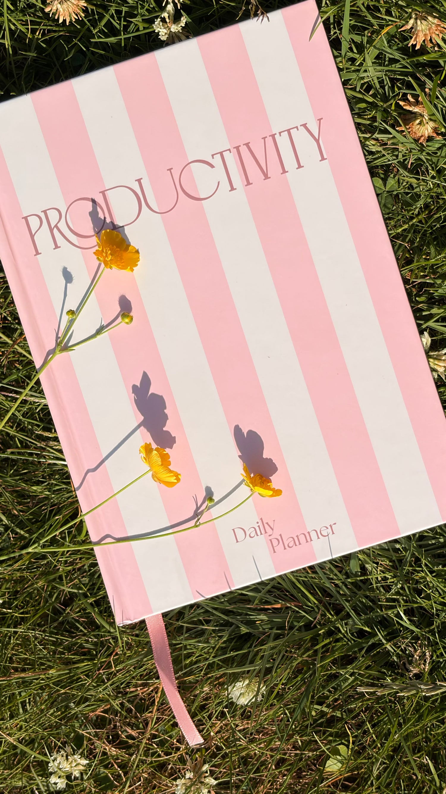 Pink and white striped daily planner with yellow flower resting on top of the planner, lying on green grass outdoors.