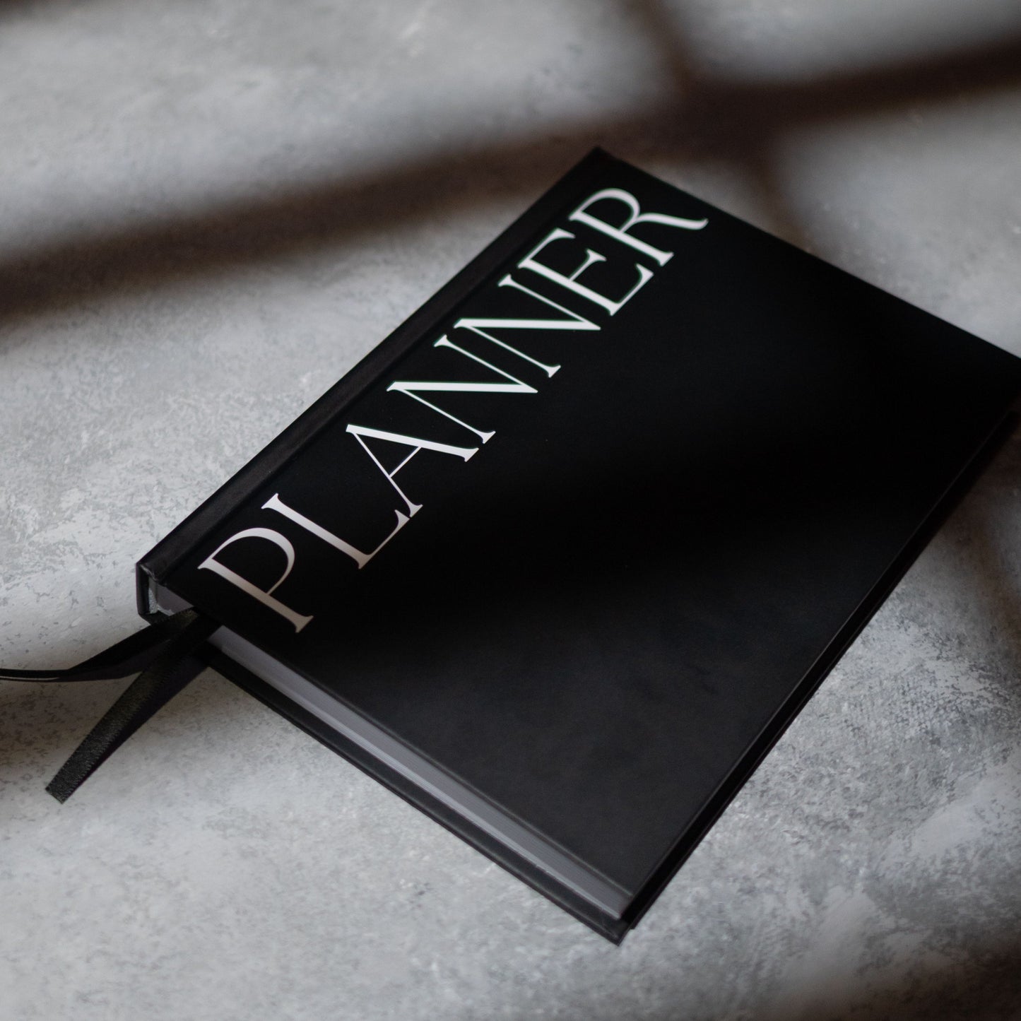A black hardcover planner with the word 'PLANNER' in large white serif letters on the cover, resting on a  grey surface with box shadows.