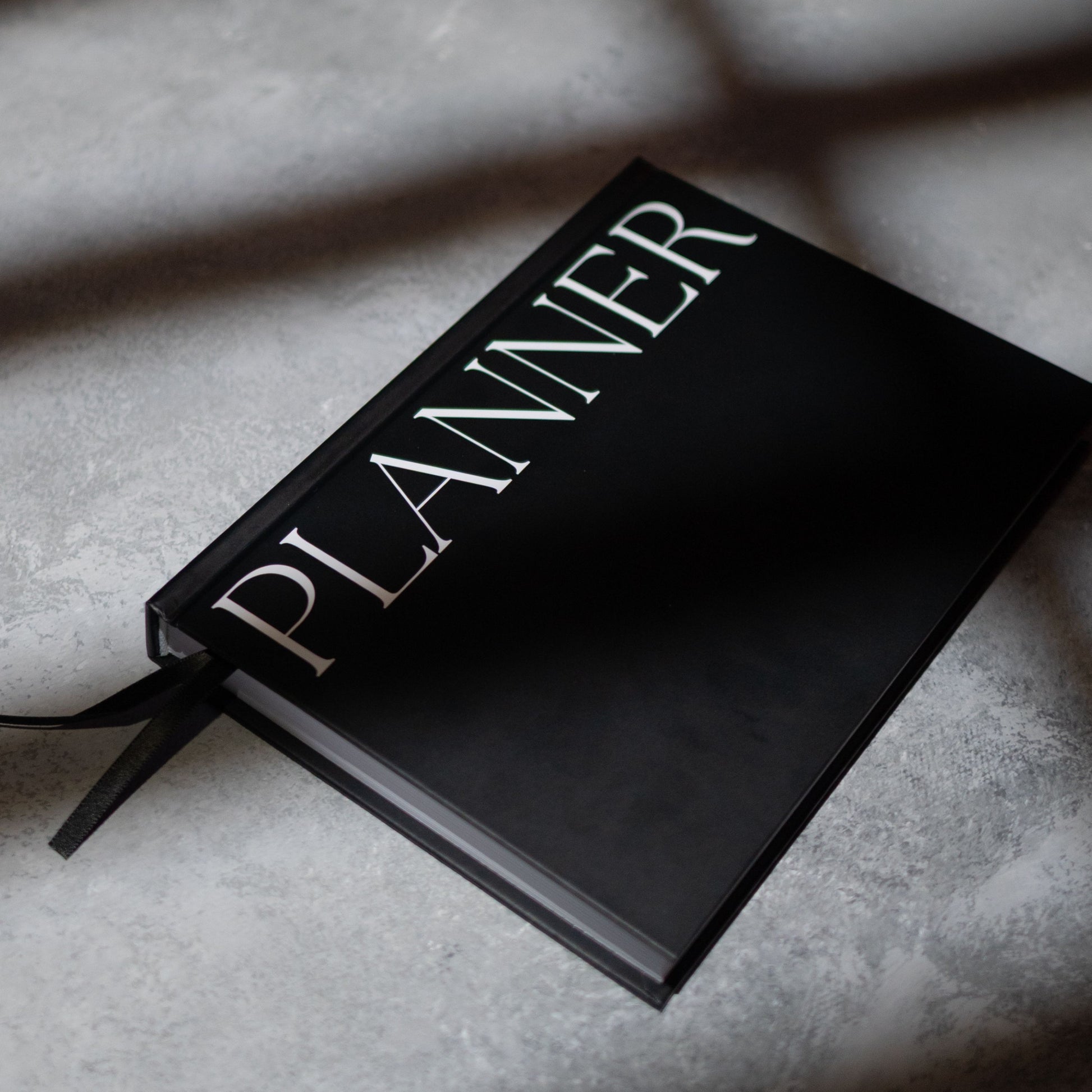 A black hardcover planner with the word 'PLANNER' in large white serif letters on the cover, resting on a  grey surface with box shadows.