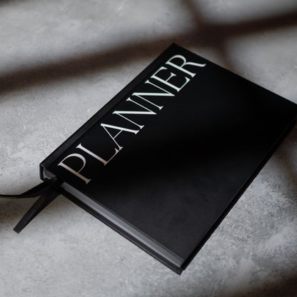 A black hardcover planner with the word 'PLANNER' in large white serif letters on the cover, resting on a  grey surface with box shadows.