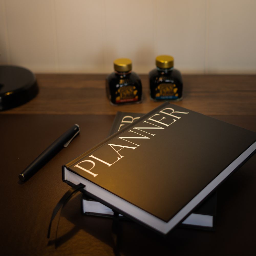 A black hardcover planner with white lettering reading 'PLANNER' sits on another planner placed on a wooden desk, with a black pen nearby and two small bottles in the background.