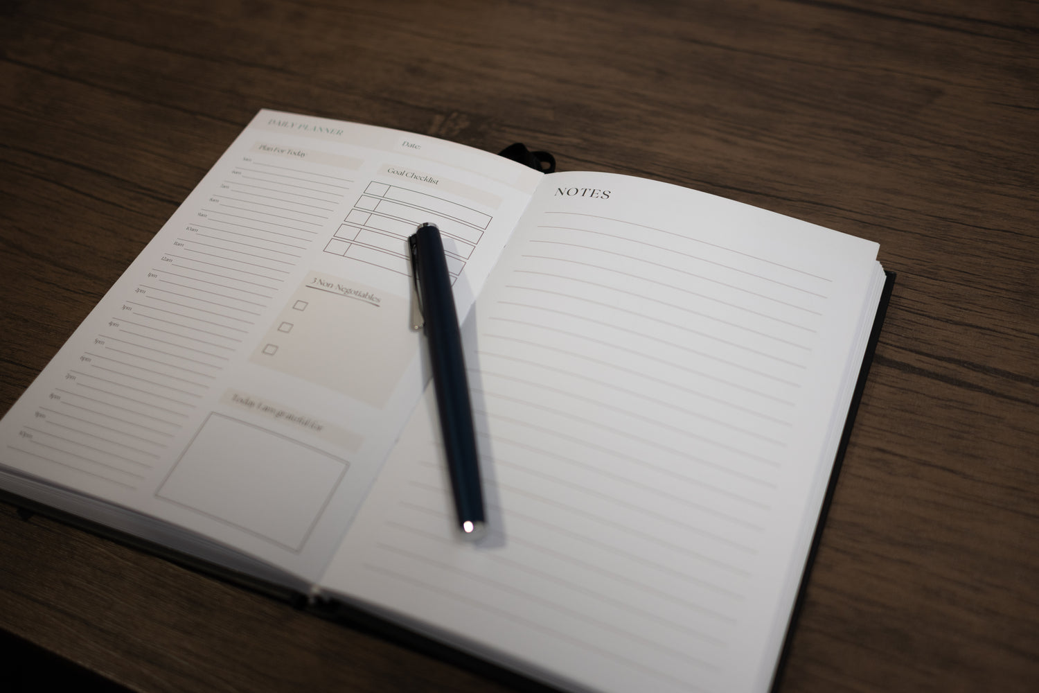 An open notebook with productivity planning framework and a lined notes page, with a black pen resting on the pages, placed on a dark wood tabletop.