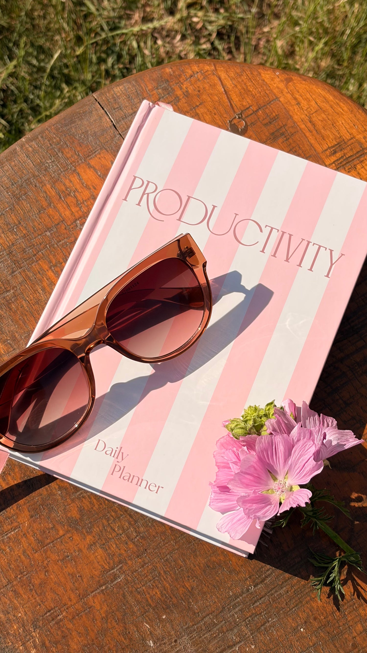 A pink and white striped daily planner with the word 'PRODUCTIVITY' on the cover, styled on a wooden table outdoors with brown sunglasses and pink flowers.