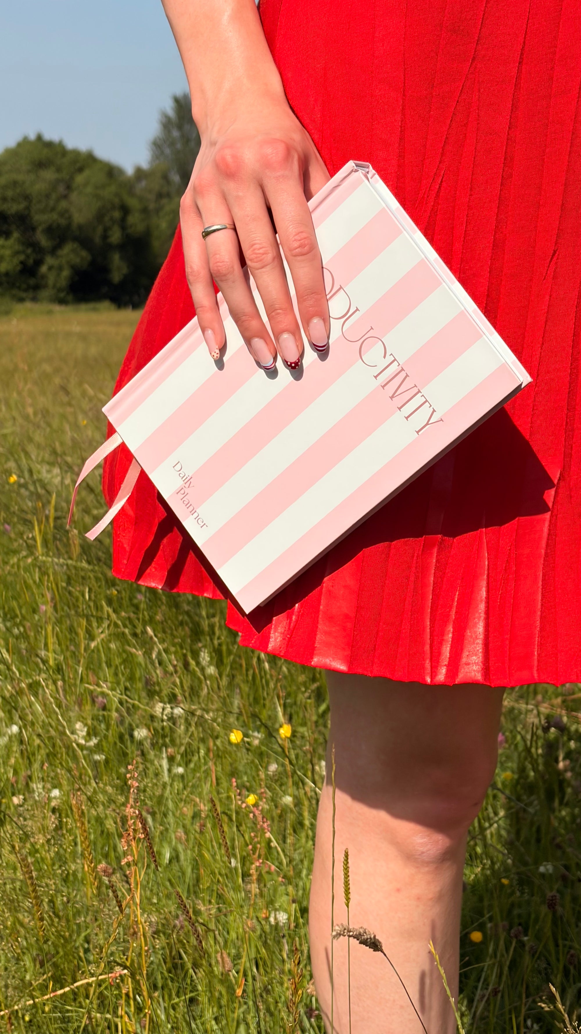 A girl wearing a bright red dress holds a pink and white striped productivity planner with the word 'PRODUCTIVITY' on the cover, standing in a field.