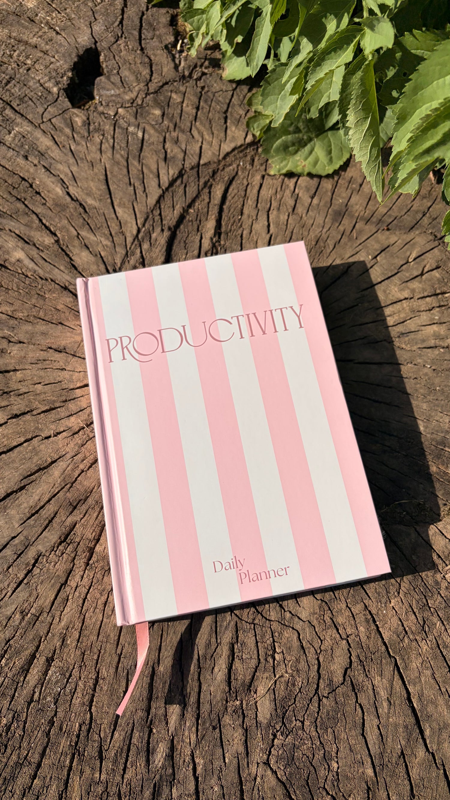 A pink and white striped hardcover journal titled 'PRODUCTIVITY' with 'Daily Planner' printed near the bottom, resting on a weathered tree stump with green leaves in the background.