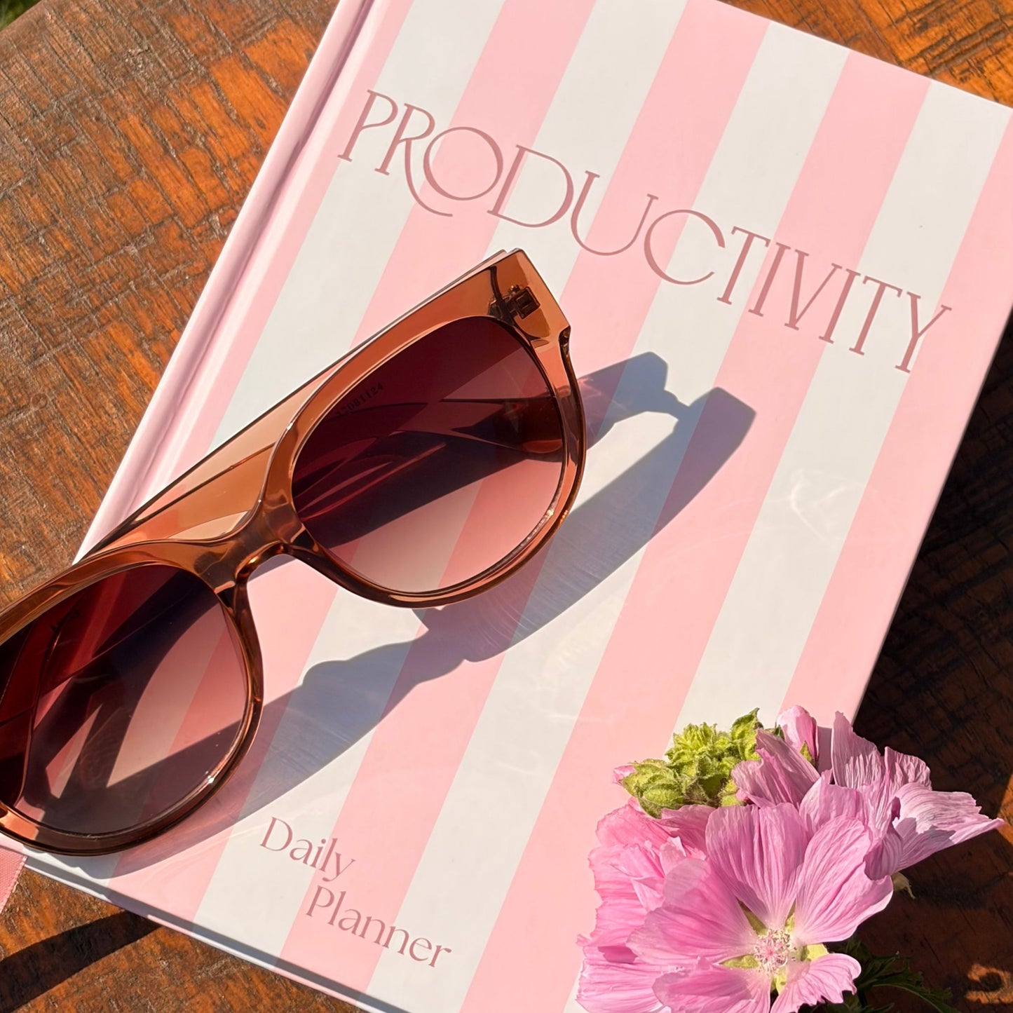 Brown sunglasses resting on a pink and white striped productivity planner titled 'Productivity Daily Planner' on a wooden table, with pink flowers in the corner.